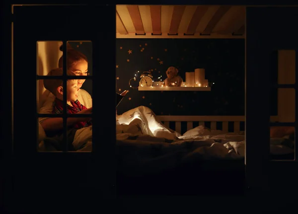 快乐的女人在家里的黑暗的卧室里 坐在舒适的床上 给快乐的男孩读着有趣的书 — 图库照片