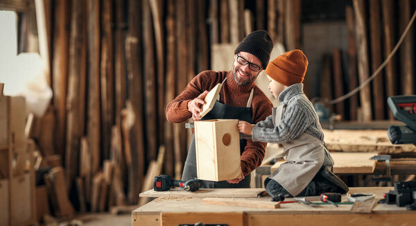Cheerful adult bearded man and little son in aprons assembling wooden bird house together while working in carpentry workshop