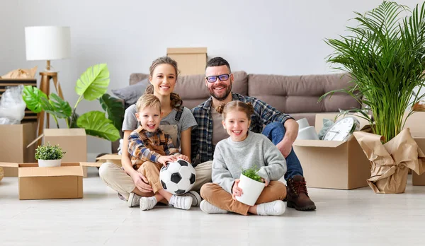 Delighted family: parents and cute kids gathering on floor in living room with unpacked boxes in new flat and looking at camera