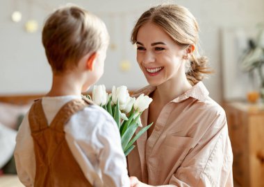 Neşeli küçük çocuğun elinde bir buket çiçekle mutlu anneye sarılırken evde tatil anneleri gününü kutlaması.