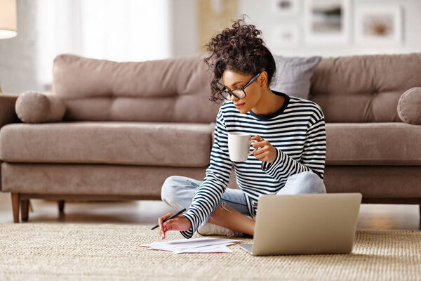 Pensive ethnic young female in glasses makes notes in documents, drinks hot coffeet and thinking while sitting cross legged near couch and working on remote project at home