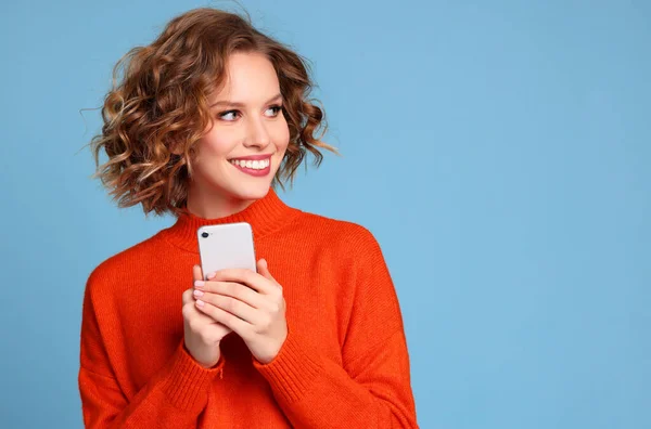 穿着时髦橙色毛衣的快乐金发女人一边笑着一边望着别处 一边拿着蓝色背景的现代智能手机 — 图库照片