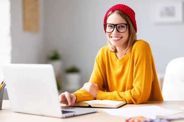 Happy young female in trendy outfit and glasses   smiling for camera while working on project in creative workplace