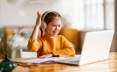 Kulaklık takmış, gülümseyen ve elini kaldırarak masada otururken online çalışmalar için dizüstü bilgisayarını kullanan sevimli bir kız öğrenci.