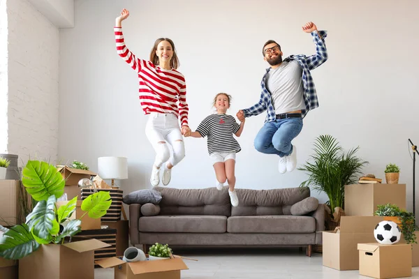 Full body delighted family: man and woman holding hands of girl and leaping up on settee near carton boxes during relocation into new apartment