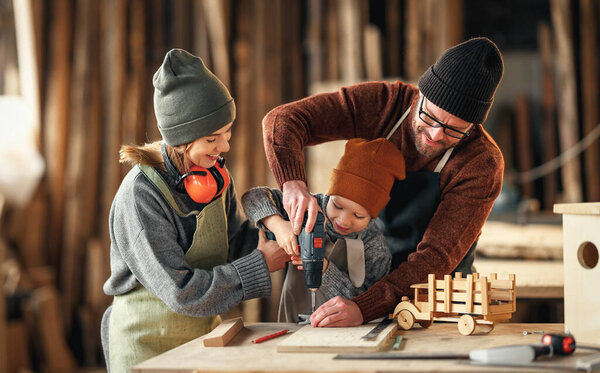 Positive mom and dad in aprons helping little son using drill while making wooden toys in carpentry workshop together