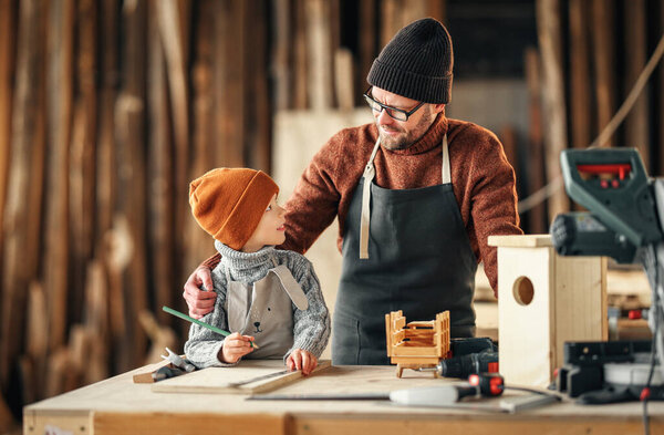 Adult bearded man in apron and little boy standing at workbench and making wooden bird house in carpentry workshop
