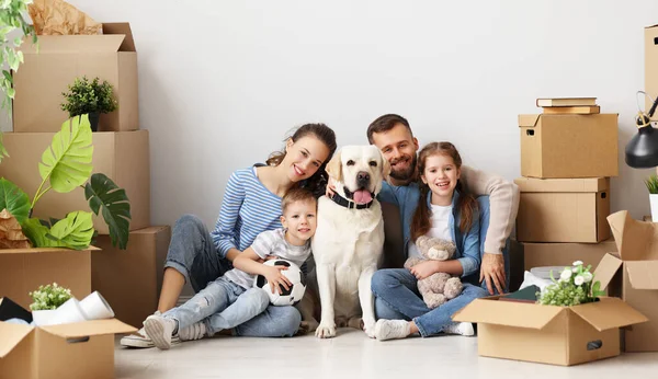 Delighted family: parents and cute kids and dog gathering on floor in living room with unpacked boxes in new flat and looking at camera