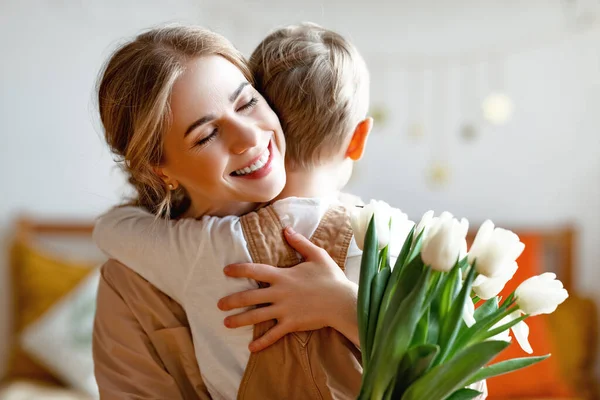 Happy woman with tulips smiling with closed eyes and embracing boy in gratitude while celebrating holiday at home