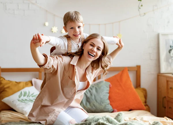 Excited mother laughing and carrying cheerful boy on back while kneeling on bed in cozy bedroom in weekend at home