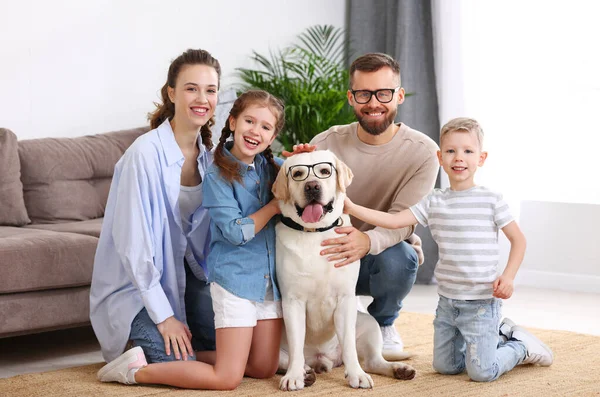 Happy parents and children gathering around adorable purebred Labrador retriever dog in funny eyeglasses and looking at camera while enjoying weekend together at home