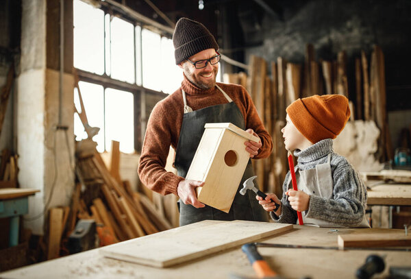Adult bearded man in apron and little boy standing at workbench and making wooden bird house in carpentry workshop