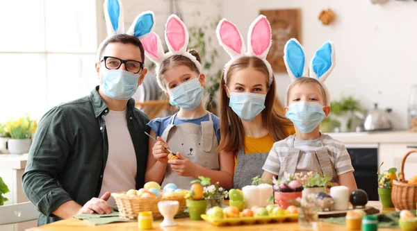 Cheerful family  in medical masks paint eggs for the holidayand  decorates house  in cozy kitchen during Easter celebration at home during the covid19 coronavirus pandemic