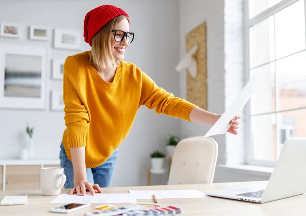 Cheerful female designer in stylish outfit and glasses smiling and drawing sketch during work on remote project at home