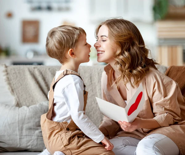 Cheerful mother hugging son and reading handmade greeting card with heart while resting on sofa during holiday celebration mothers day at home