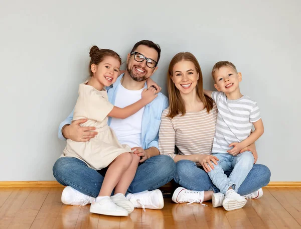 Full body of cheerful family with little kids looking at camera while sitting together on floor near wall in empty room of new home