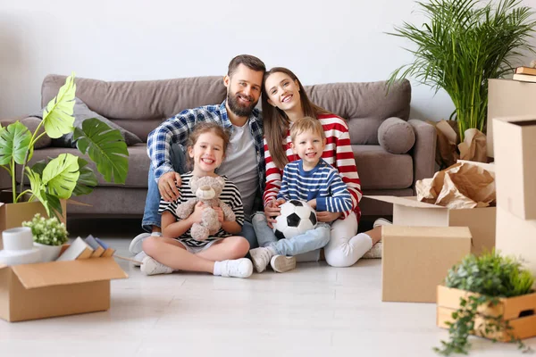 Delighted parents and cute kids gathering on floor in living room with unpacked boxes in new flat and looking at camera