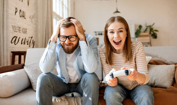 Frustrated bearded man touching head and watching excited female playing videogame while resting on couch in weekend at home together