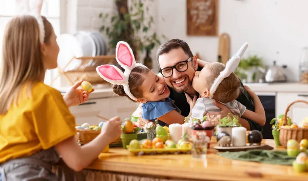Loving cute children wearing funny bunny ears headbands embracing and kissing cheerful young father while gathering with family at table in kitchen for Easter eggs painting