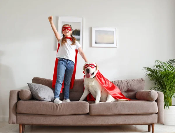 全身上下都是穿着红色超级英雄斗篷 戴着面具的小女孩 一边举手表决 一边在家里客厅里与穿着类似服装的滑稽狗狗玩耍 — 图库照片