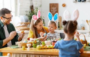 Tavşan kulaklığı takan neşeli aile modern aydınlık mutfakta toplanıyor ve Paskalya yumurtalarını birlikte acıtıyorlar.