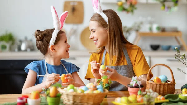 快乐的年轻妈妈和可爱的女儿戴着滑稽的兔子耳朵帽带 在厨房里一起画复活节彩蛋 面带微笑地互相看着对方 — 图库照片