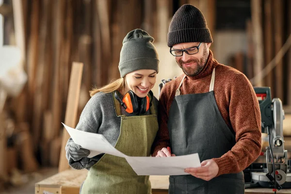 Positive male and female craft small business owners in aprons discussing paper draft while working together in carpentry workshop