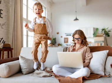 Küçük oğlu eğlenirken, zıplarken ve gürültü yaparken genç anne kanepede oturup dizüstü bilgisayarla çalışıyor.
