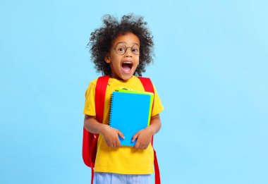 Afro saçlı, sıradan giyinmiş ve gözlüklü sevimli, çalışkan küçük siyah okul çocuğu, mavi arka planda defterlerle dururken hayretle çığlık atıyordu.
