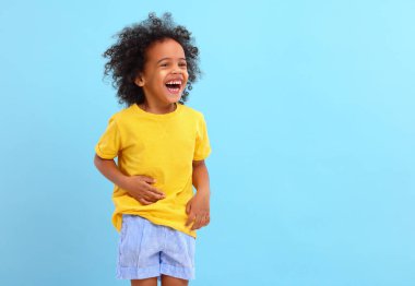 Afro saçlı keyifli küçük zenci çocuk günlük kıyafetlerle gülüyor ve mavi arka plana bakıyor.