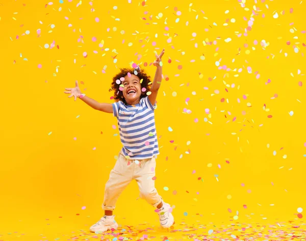 黄色の背景にカラフルなコンフェッティをキャッチしようとしている間 スタイリッシュな服の中でアフロの髪と喜びの小さな黒い子供の完全な体笑いとジャンプ — ストック写真