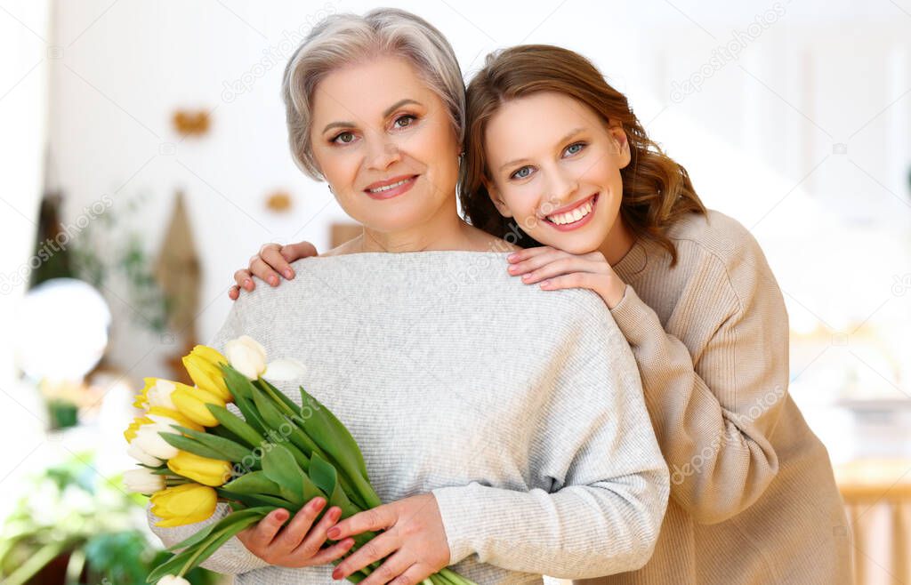 Positive elegant elderly woman with bunch of fresh tulips smiling and looking at camera while standing at home with happy young daughter embracing shoulders gently