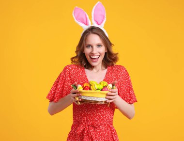 Neşeli, kırmızı elbiseli, tavşan kulaklı, gülümseyen ve kameraya bakan, sarı arka planda renkli Paskalya yumurtalarının olduğu sepetle duran genç bir kadın.