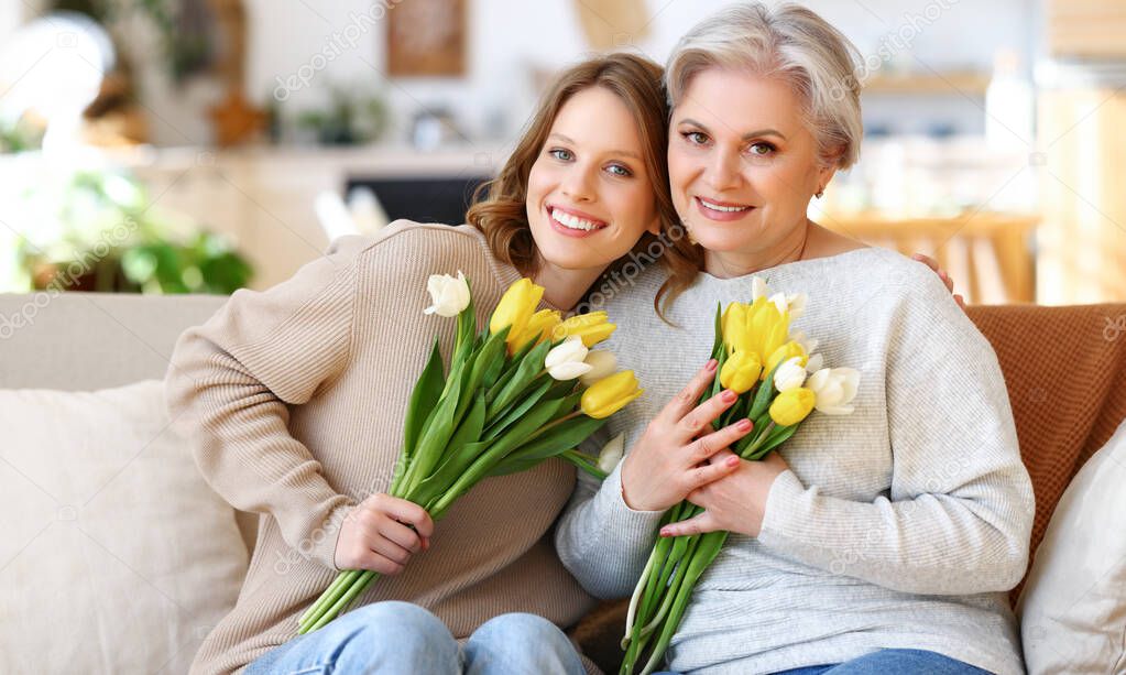 Positive young daughter with senior mother in casual clothes smiling and hugging while sitting on sofa at home with bunches of fresh delicate tulips during celebration of international Women Day