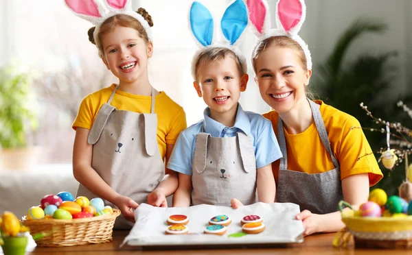 快乐的母亲和可爱的兄弟姐妹们 戴着小兔子耳朵 带着手工制作的家庭饼干聚集在餐桌旁 准备在家里庆祝复活节 — 图库照片