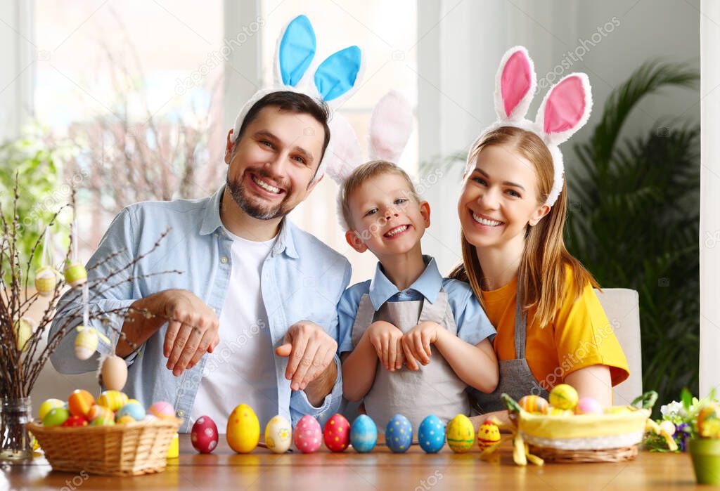Happy parents and adorable boy wearing cute bunny ears sitting together at table with colorful painted eggs while preparing for Easter holiday and looking at camera