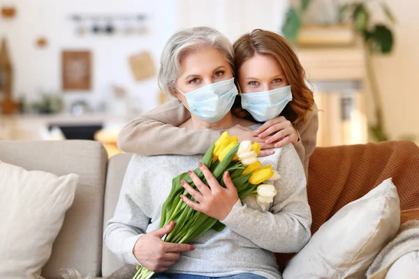 Anonymous young woman in face medical mask embracing senior mother while congratulating International Mothers Day and giving bouquet of fresh tulips at home