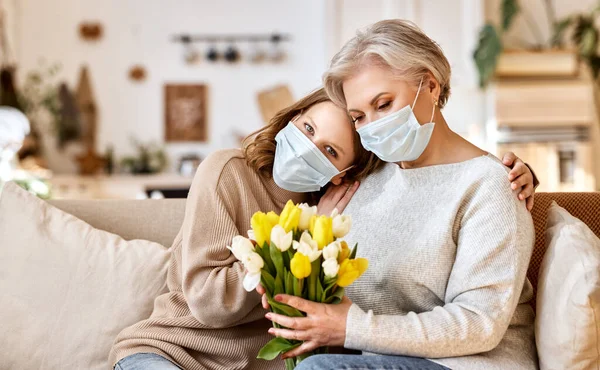 Anonymous young woman in face mask embracing senior mother while congratulating International Mothers Day and giving bouquet of fresh tulips at home