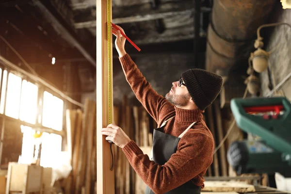 Focused adult male woodworker in apron and knitted gat cutting wooden plank while working in carpentry workshop