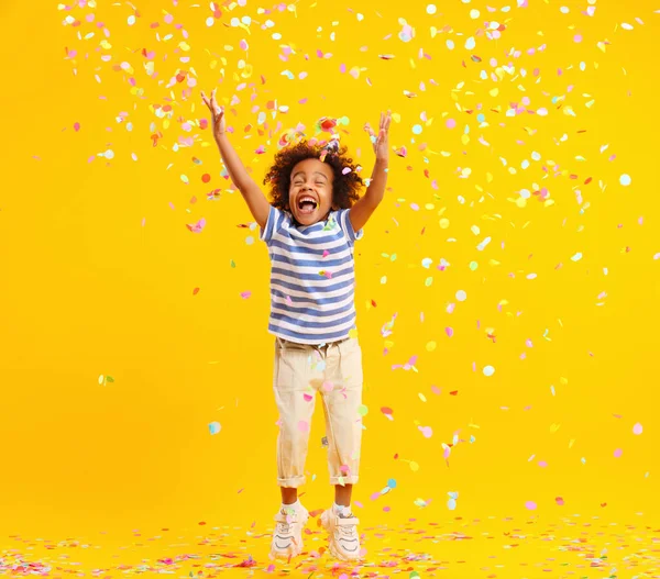 黄色の背景に対する誕生日のお祝いの間にカラフルなコンフェッティをキャッチしようとしている間 アフロの髪の笑いとジャンプと楽しい小さな黒い子供 — ストック写真