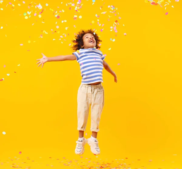 黄色の背景に対する誕生日のお祝いの間にカラフルなコンフェッティをキャッチしようとしている間 アフロの髪の笑いとジャンプと楽しい小さな黒い子供 — ストック写真