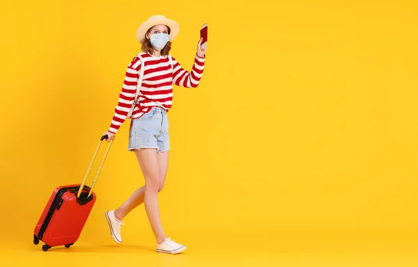 カジュアルな服を着た認識できない若い旅行者の全身と スーツケース付きの黄色の背景を歩くチケットとパスポート付きのフェイスマスク — ストック写真