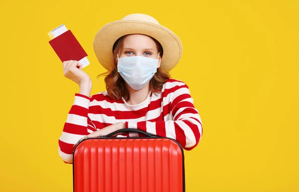 カジュアルな服の帽子と顔のマスクを手にパスポートとチケットを保持し 黄色の背景に幸せとカメラを見てスーツケースと興奮した若い女性観光客 — ストック写真