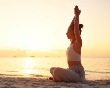 Lotus 'ta kumlu sahilde oturmuş Namaste' nin elleri yukarıda ve gün batımına karşı meditasyon yaparken uzun ve sakin bir kadın.