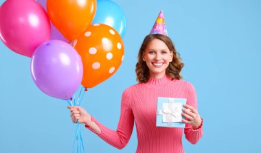 Neşeli genç bir bayan gülümseyip kameraya bakarken mavi arka planda renkli balonlar ve paketlenmiş doğum günü hediyeleri gösteriyor.