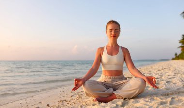Spor kıyafetleri içinde uzun boylu sakin bir kadın Lotus 'ta kumlu sahilde oturuyor ve gün batımına karşı meditasyon yapıyor.