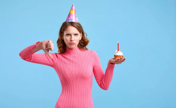 不幸な若いです女性ともに誕生日カップケーキ見ますカメラとジェスチャー親指ダウンでお祝いの間に青の背景 — ストック写真