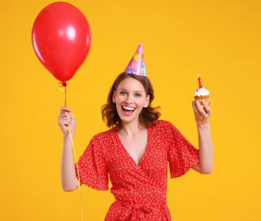 Optimistic female with red balloon and birthday cupcake  smiling and looking at camera against blue background