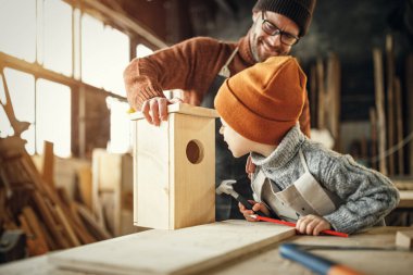 Neşeli yetişkin sakallı adam ve küçük oğlu marangozluk atölyesinde çalışırken ahşap kuş evini birleştiriyorlar.