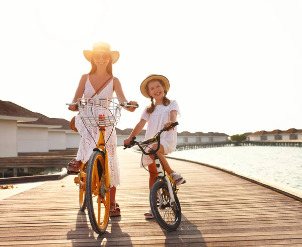 Full body of cheerful family: young woman and little daughter in white summer dresses and hats smiling  and looking at camera while riding bicycles on pier near sea